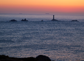 Sunset longships This landscape photograph captures the Longships lighthouse positioned off the coast of Cornwall in the United Kingdom during an evening sunset in early spring. The image displays the striking contrast between the orange and pink hues of the sky and the deep blue tones of the sea, showcasing the natural beauty of the Cornish coastline. The Longships lighthouse stands prominently on the islet, surrounded by rocky outcrops emerging from the water. In the distance, cargo boats are visible on the horizon, highlighting maritime activity in the region. The photograph exemplifies the tranquil nature of the scene, with elements of both the rugged coastline and the open sea under the atmospheric light of a spring evening sunset.
